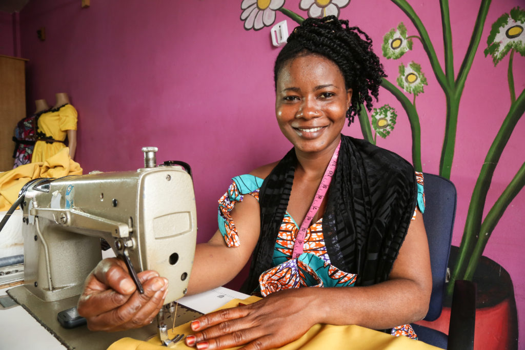Faiza, a young woman from Ghana sits proudly with her sewing machine against a brightly coloured pink wall.