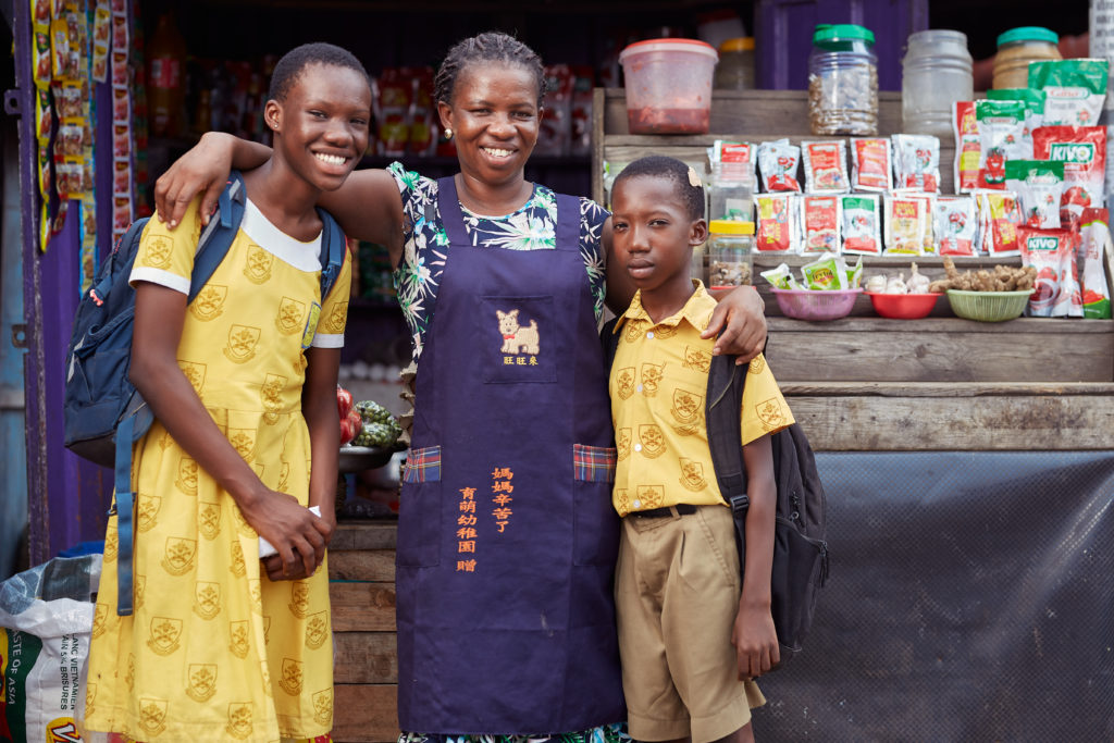 Josephine, a widow and mother of four, stands with two of her children looking happy in front of her business stall in the market.