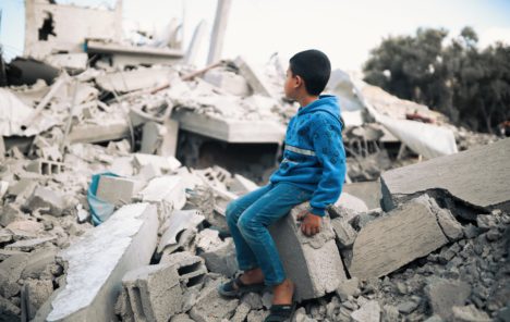 A boy in blue sits on rubble staring into the distance at the destroyed buildings