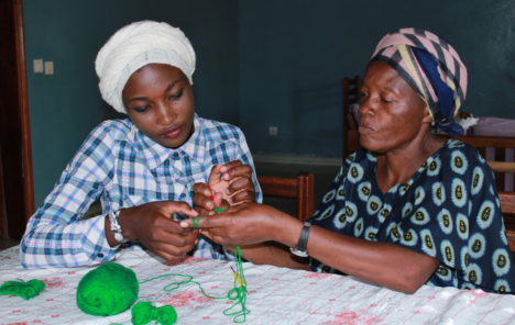 Jeanne (left) is being taught by an older lady how to knit. They sit at a table together holding the green wool and concentrating on their task