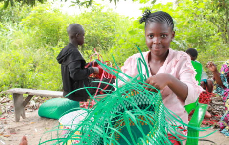 A young person sits outdoors smiling while holding a large, half-finished basket made from green fibre.