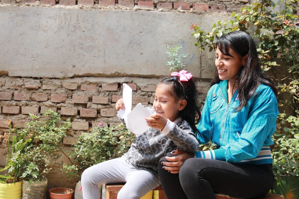 A mother and her young daughter sit outside in front of a brick wall, laughing as the young girl lifts two pieces of paper up into the air to see what is on them.